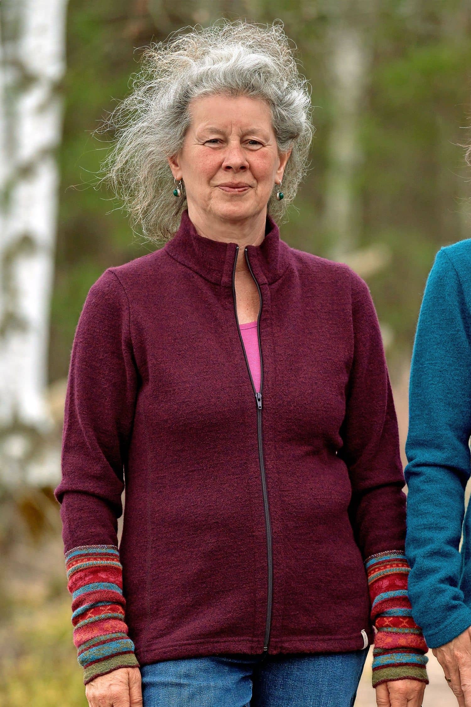 Frau steht im Wald und trägt Jeans, pinkes Shirt, beerefarbene Jacke und gemusterte Armstulpen