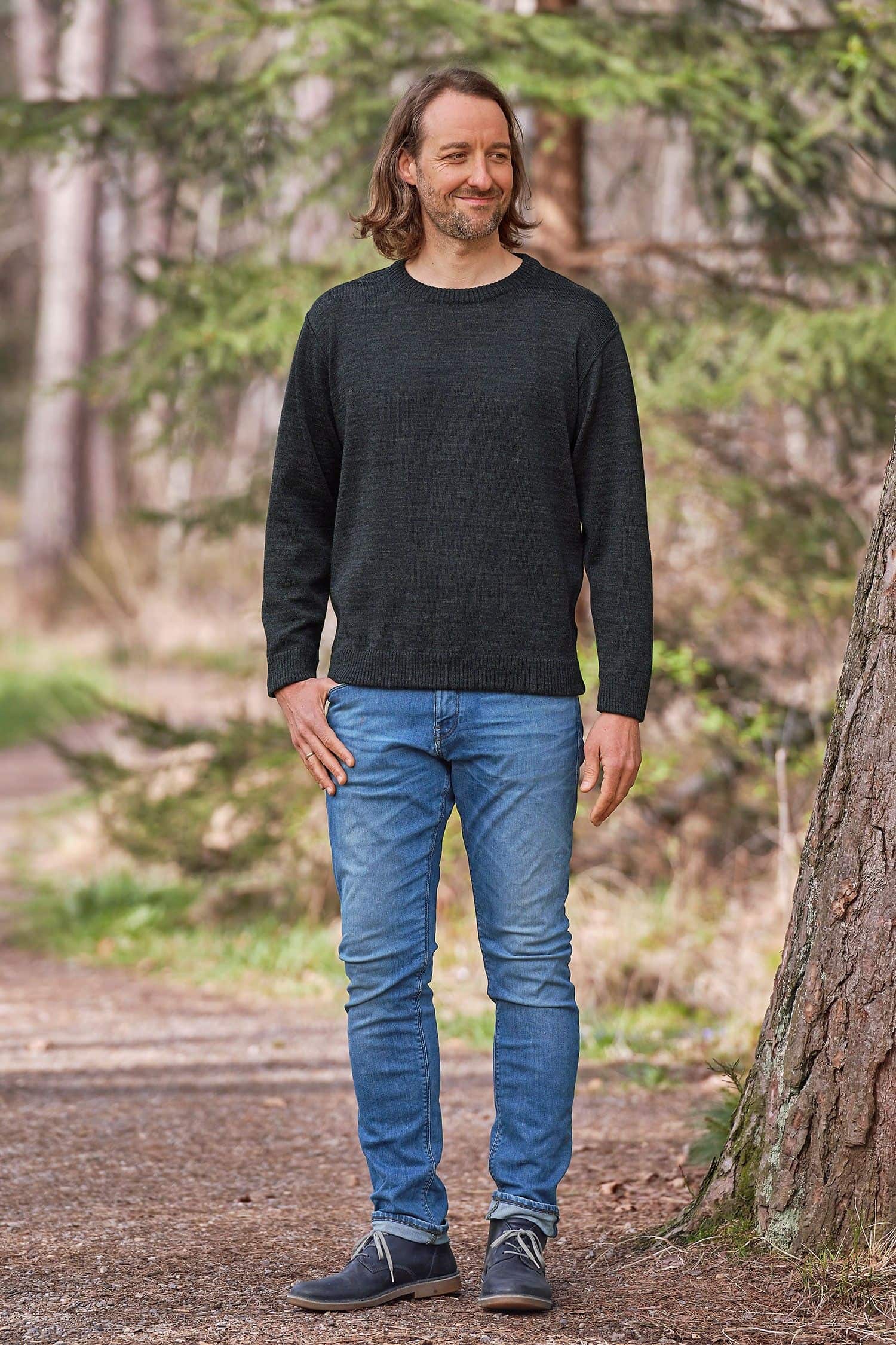 Mann steht mit Jeans und anthrazitgrauem Rundhalspullover aus bio Wolle (kbT) vom Finkhof neben einem Baum