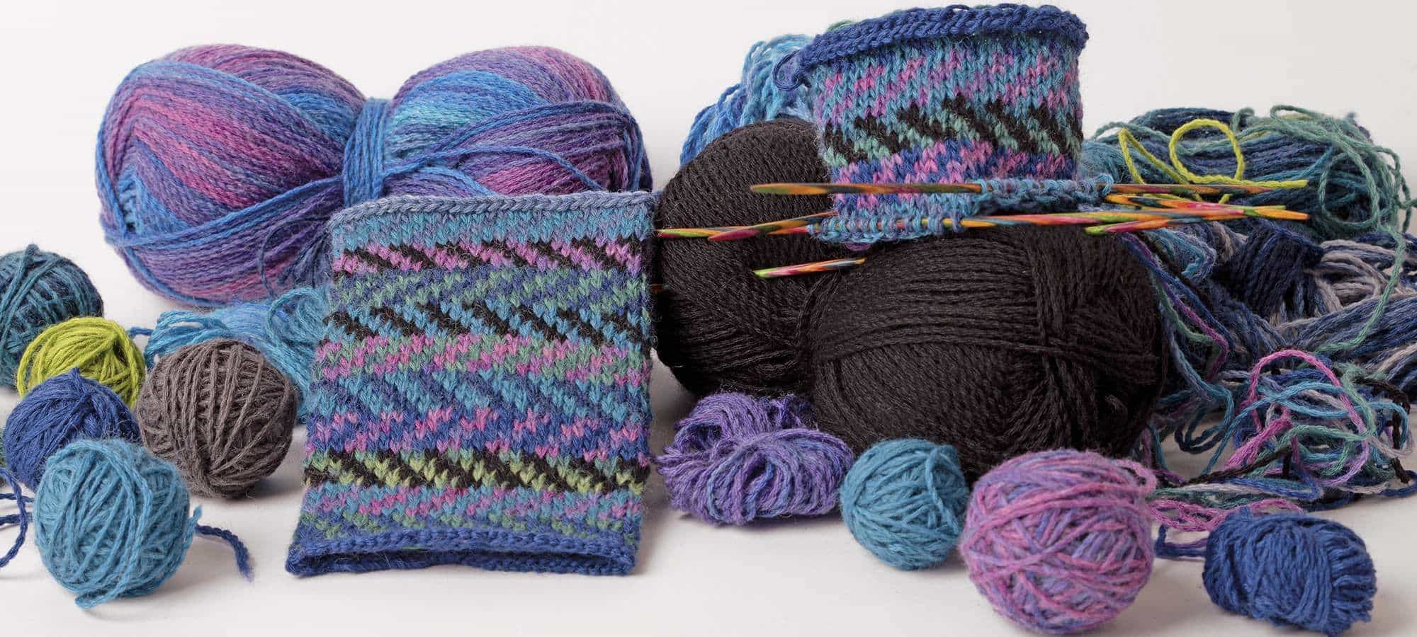 bunt gemusterte Pulswärmer mit Wollen in lila und Blautönen auf weißem Untergrund als Banner für Strickanleitung