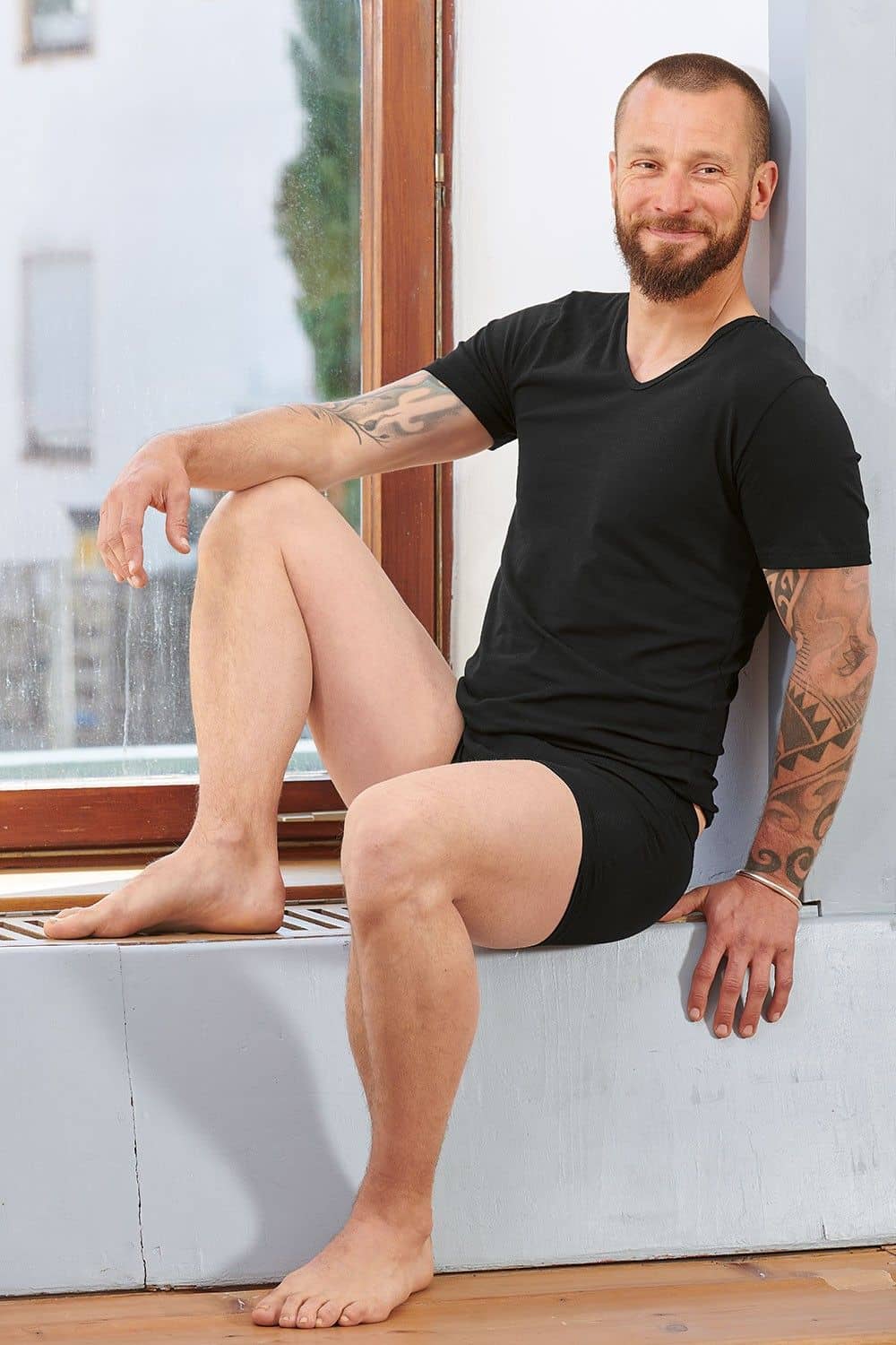 Mann sitzt auf Fensterbank und trägt Boxershorts und schwarzes Kurzarmshirt aus bio Baumwolle (kbA) und Elasthan