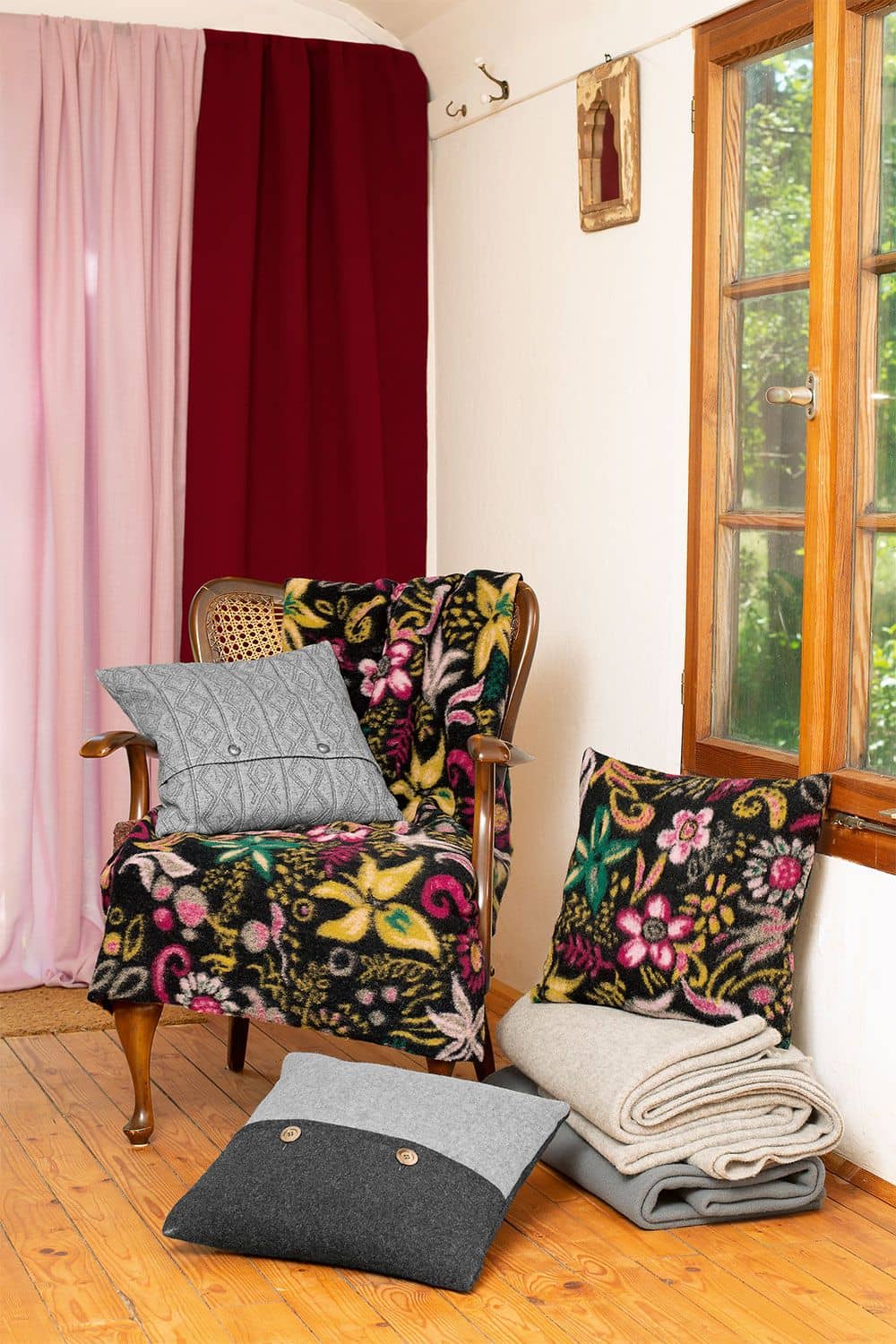 eingerichtete Wohnzimmerecke vor Fenster mit Korbstuhl und Decken und Kissen aus floralem Walkstoff