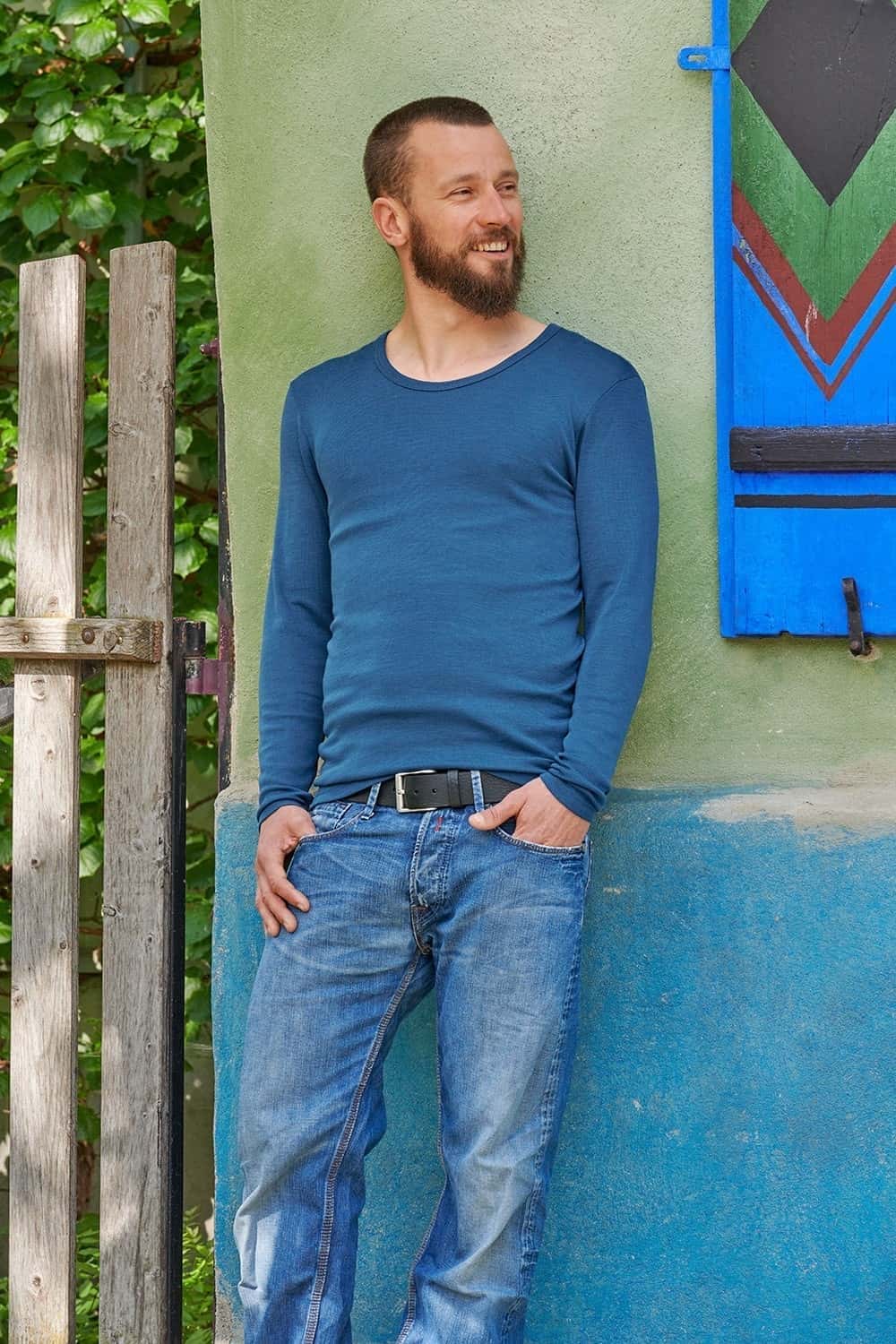 Mann lehnt an grüner Wand und trägt ein dunkelblaues Langarmshirt aus bio Wolle (kbT) und Seide und eine Jeans