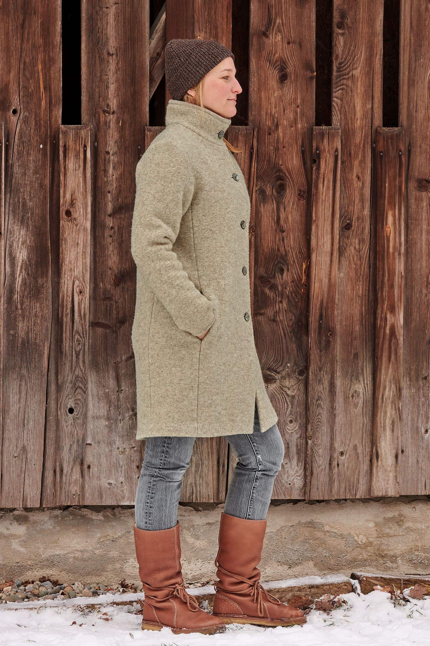 Frau steht vor Holzwand und trägt braune Mütze und einen grauen Finkhof Wollwalkmantel aus bio Wolle (kbT)