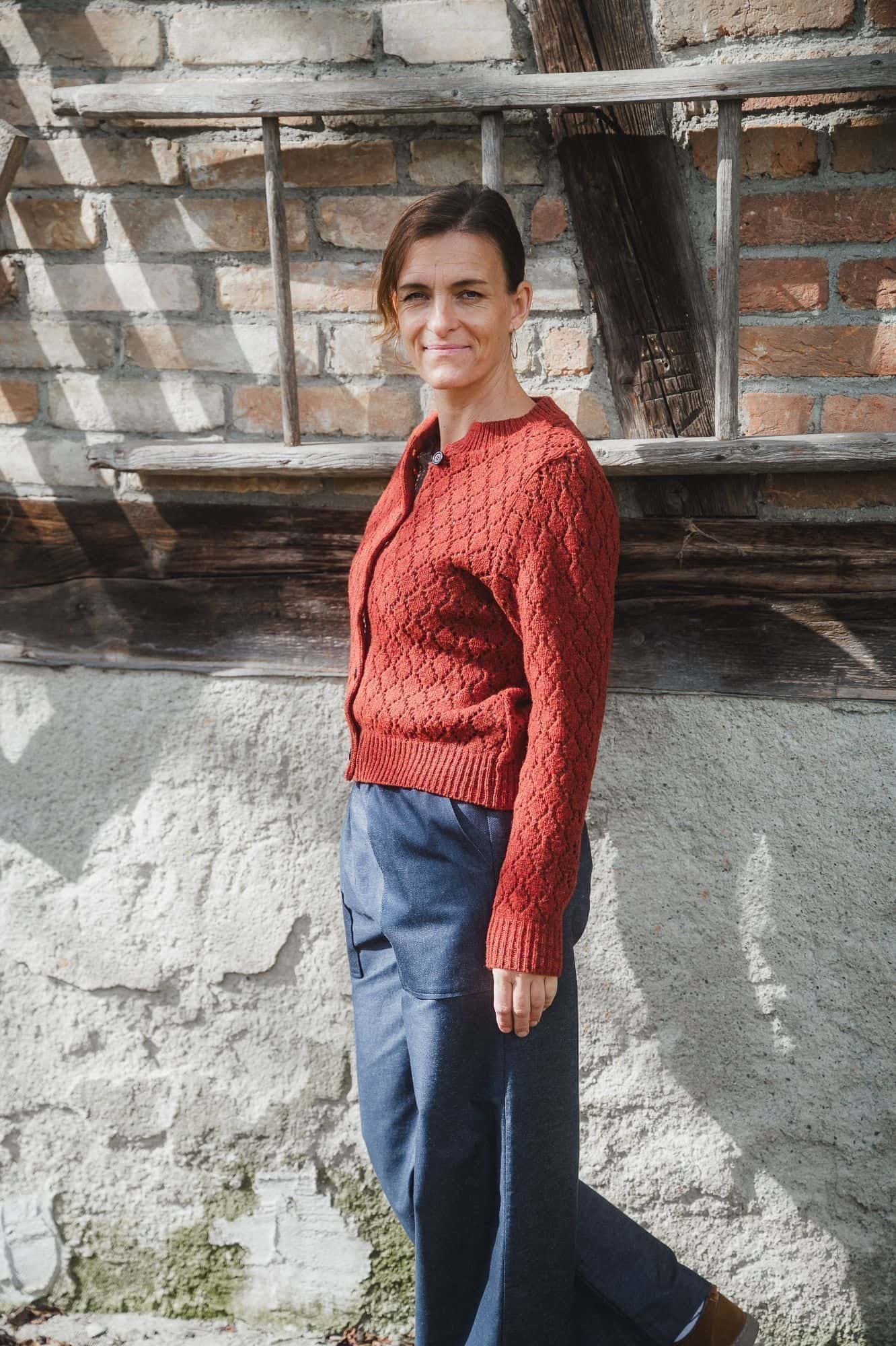 Finkhof-Ajour-Damen-Strickjacke siena Gr. S Frau trägt eine weite blaue Hose und eine rot orange Strickjacke mit Ajourmuster aus Bio-Wolle vom Finkhof