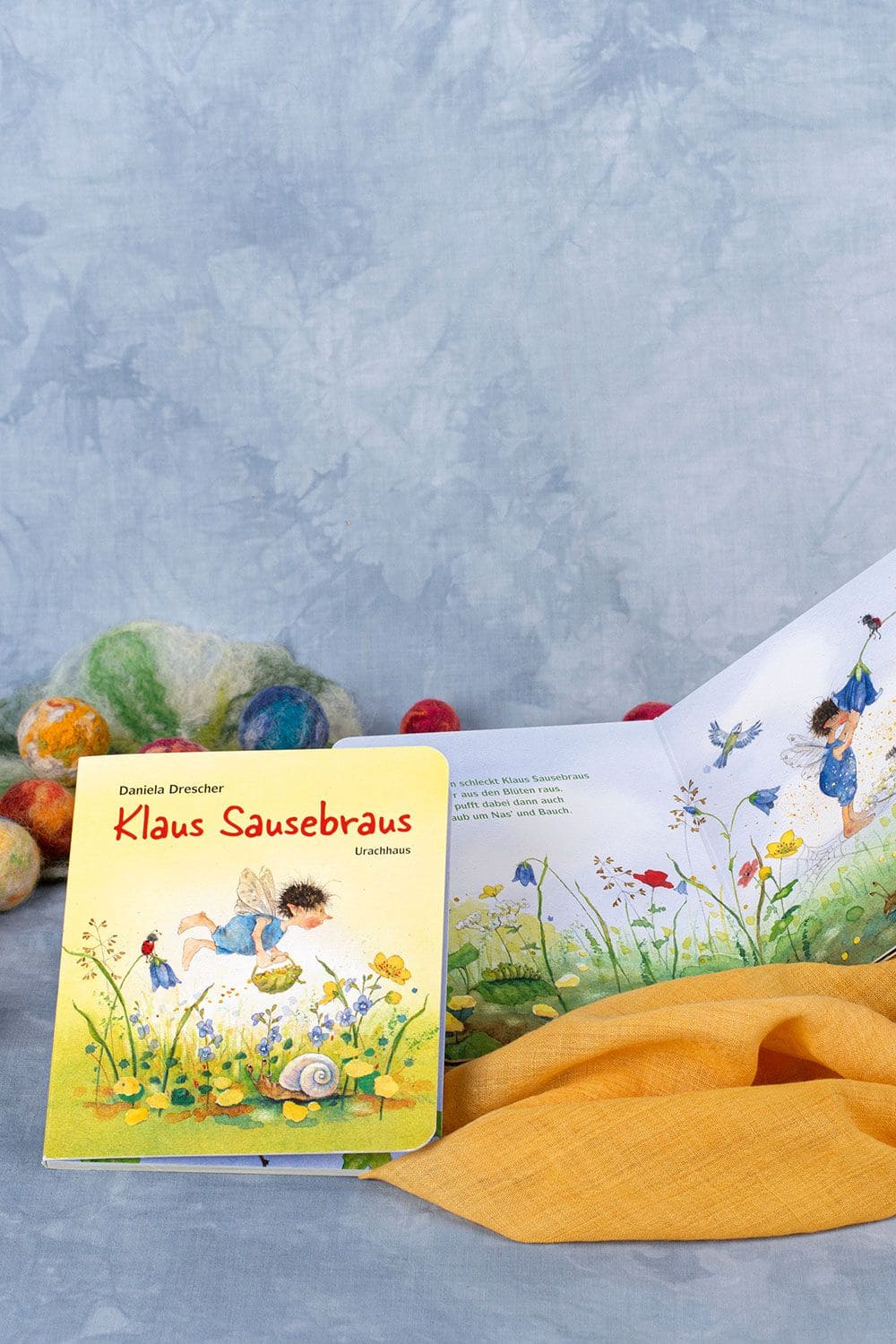 Klaus Sausebraus Buch ,,Klaus Sausebraus" auf blauem Untergrund mit aufgeschlagenem Exemplar an der Seite