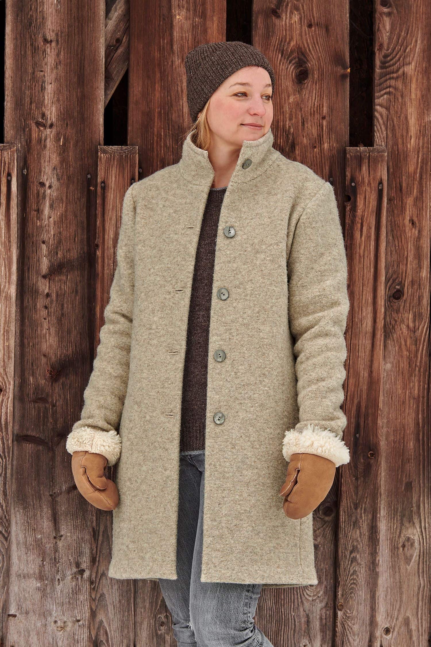 Frau steht vor Holzwand und trägt braune Mütze, Handschuhe und einen grauen Wollwalkmantel aus bio Wolle (kbT) vom Finkhof