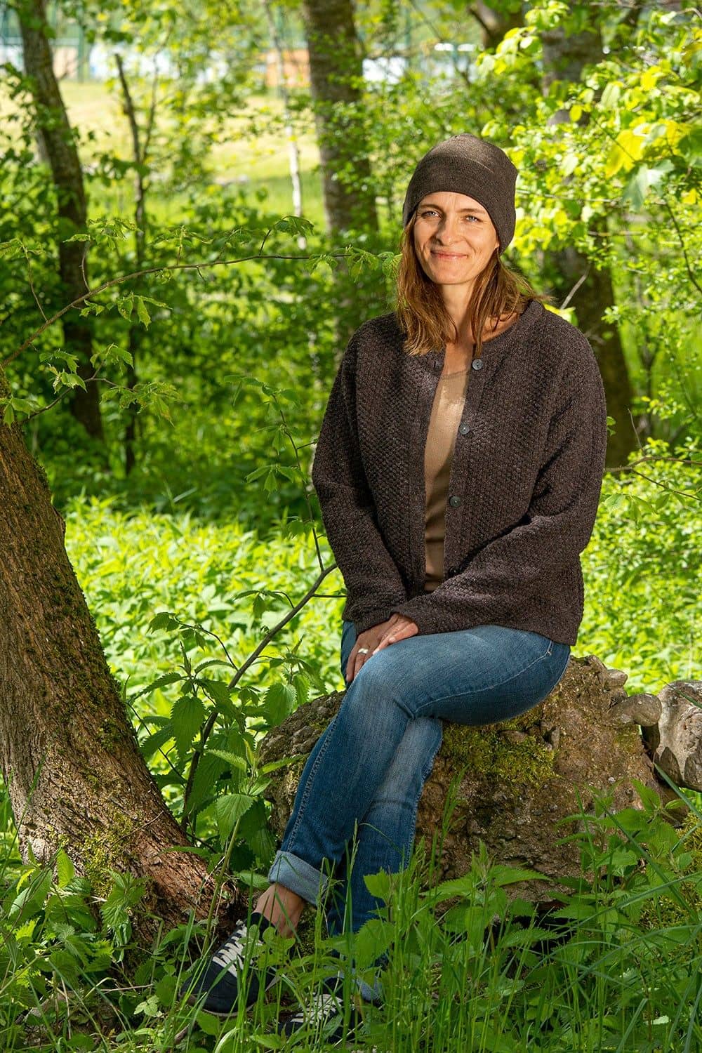 Frau sitzt auf Stein und trägt Jeans, braune Mütze und braune Strukturjacke vom Finkhof aus bio Wolle (kbT)