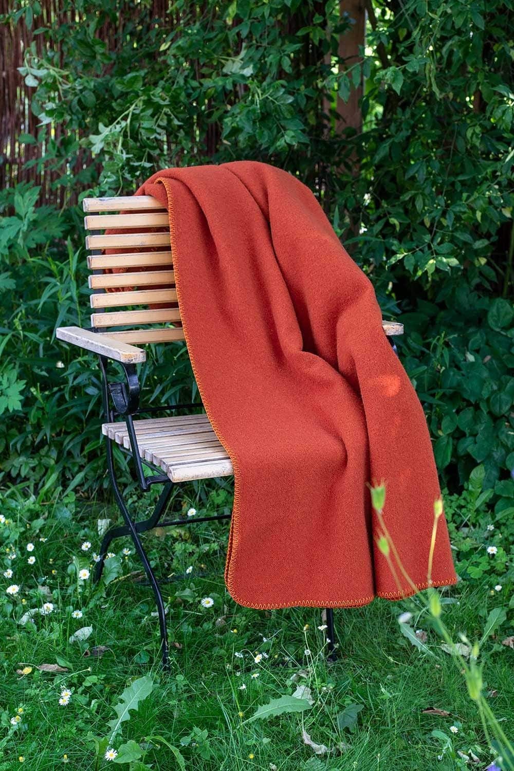 rostrote Wolldecke mit oranger Kante aus bio Wolle (kbT) vom Finkhof hängt über einem Gartenstuhl
