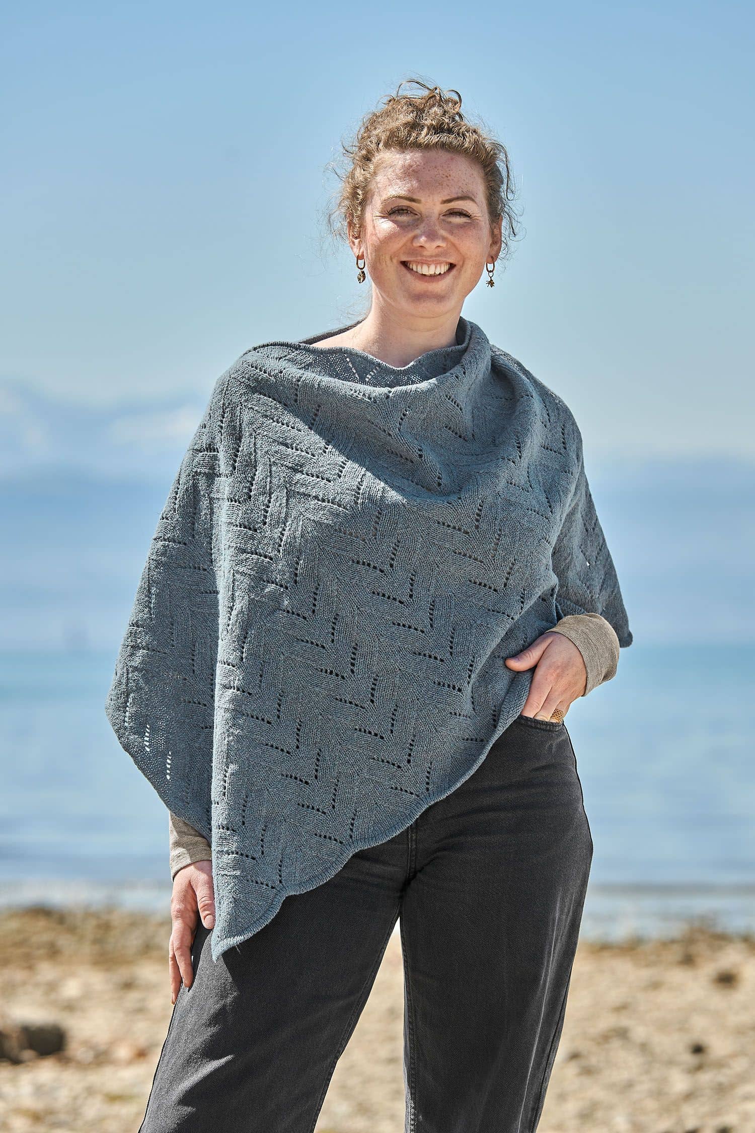 Finkhof-Ajour-Poncho Frau trägt eine schwarze Jeans und einen rauchblauen Poncho mit Ajour Muster vom Finkhof aus bio Merinowolle (kbT)