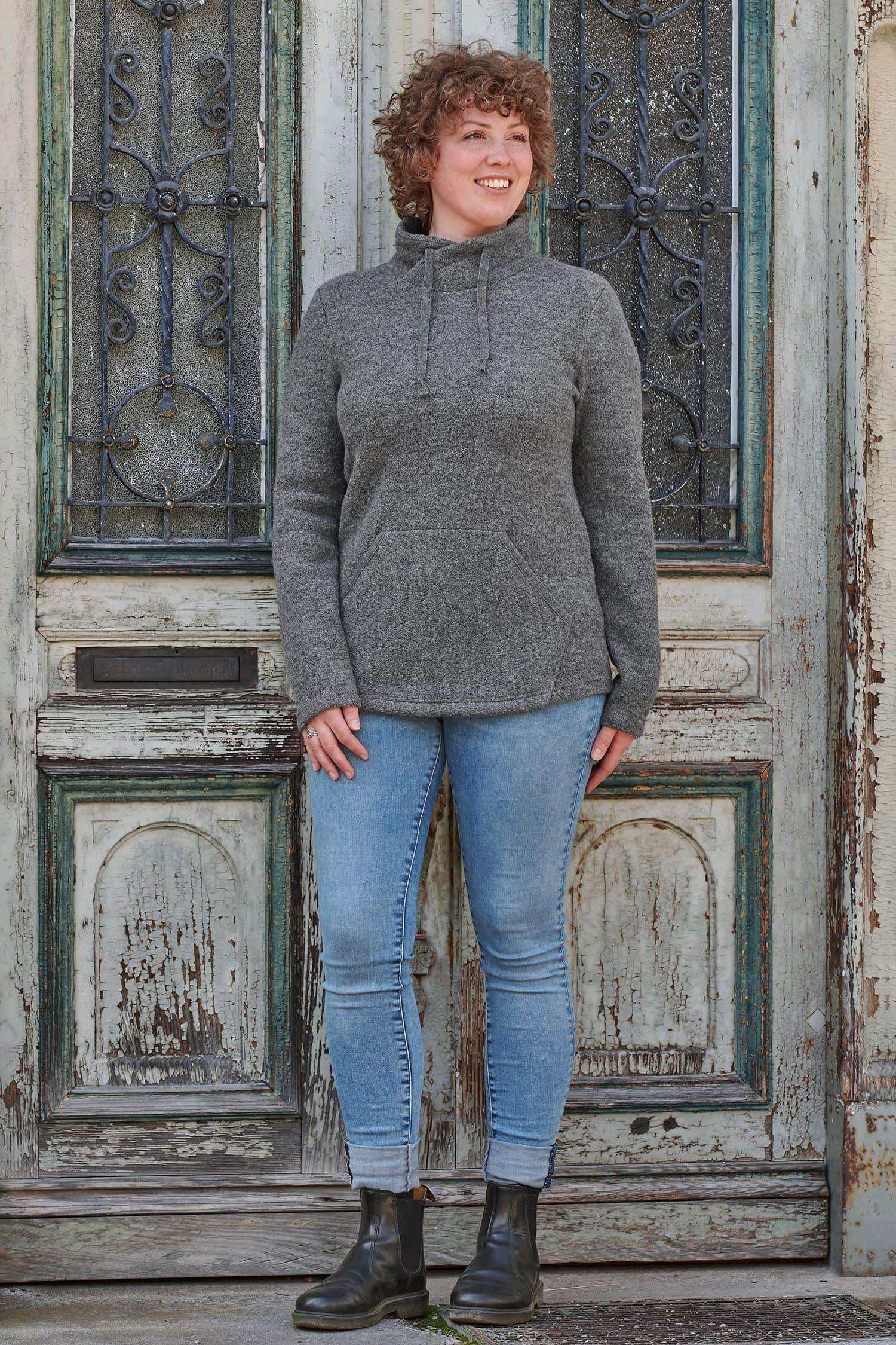 Frau trägt grauen Pullover aus bio Wolle (kbT) mit Jeans und steht vor alter blauer Holztür
