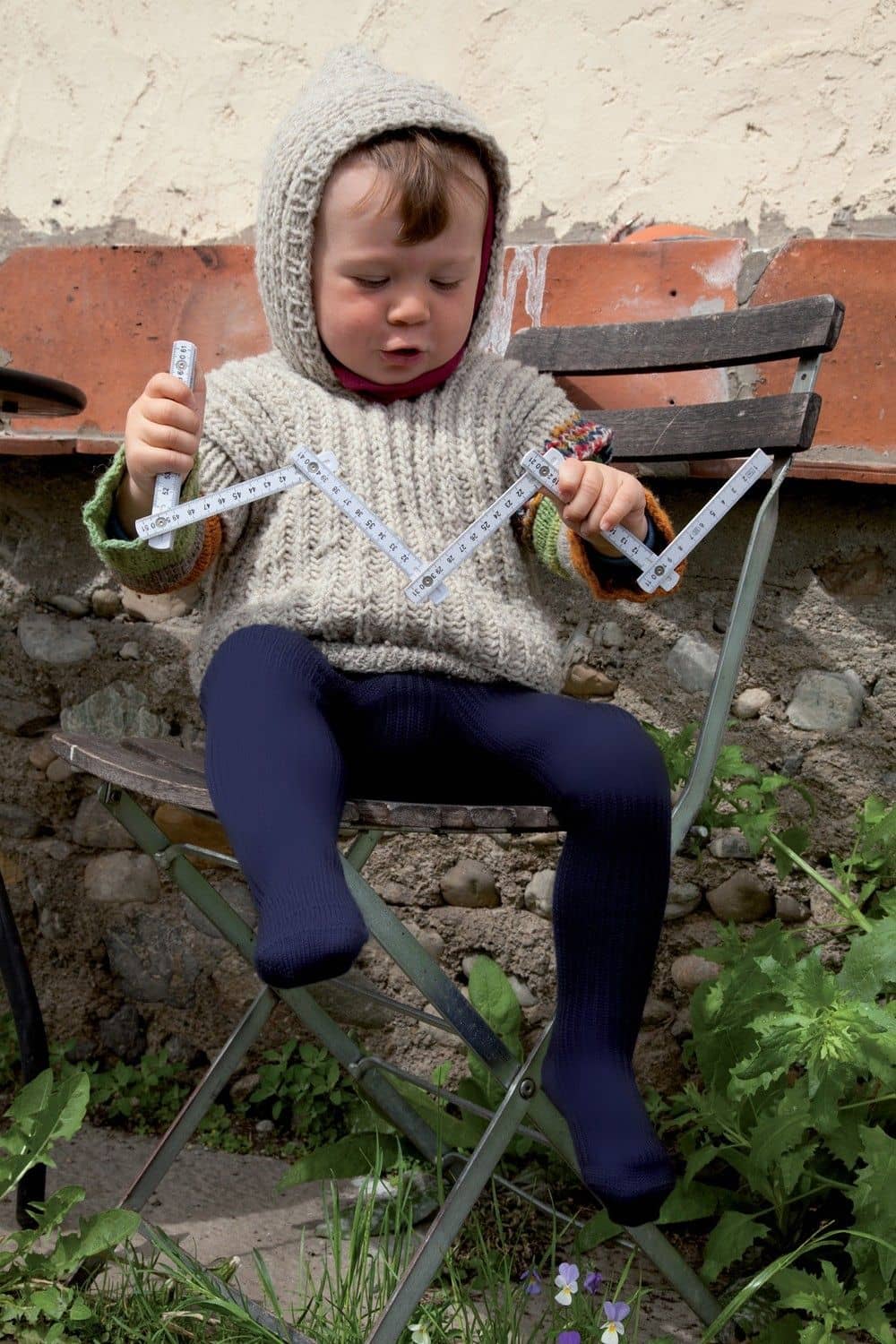 Kind mit Wollpulli und Meterstab sitzt auf einem Stuhl und trägt eine marineblaue Wollstrumpfhose aus bio Wolle (kbT)