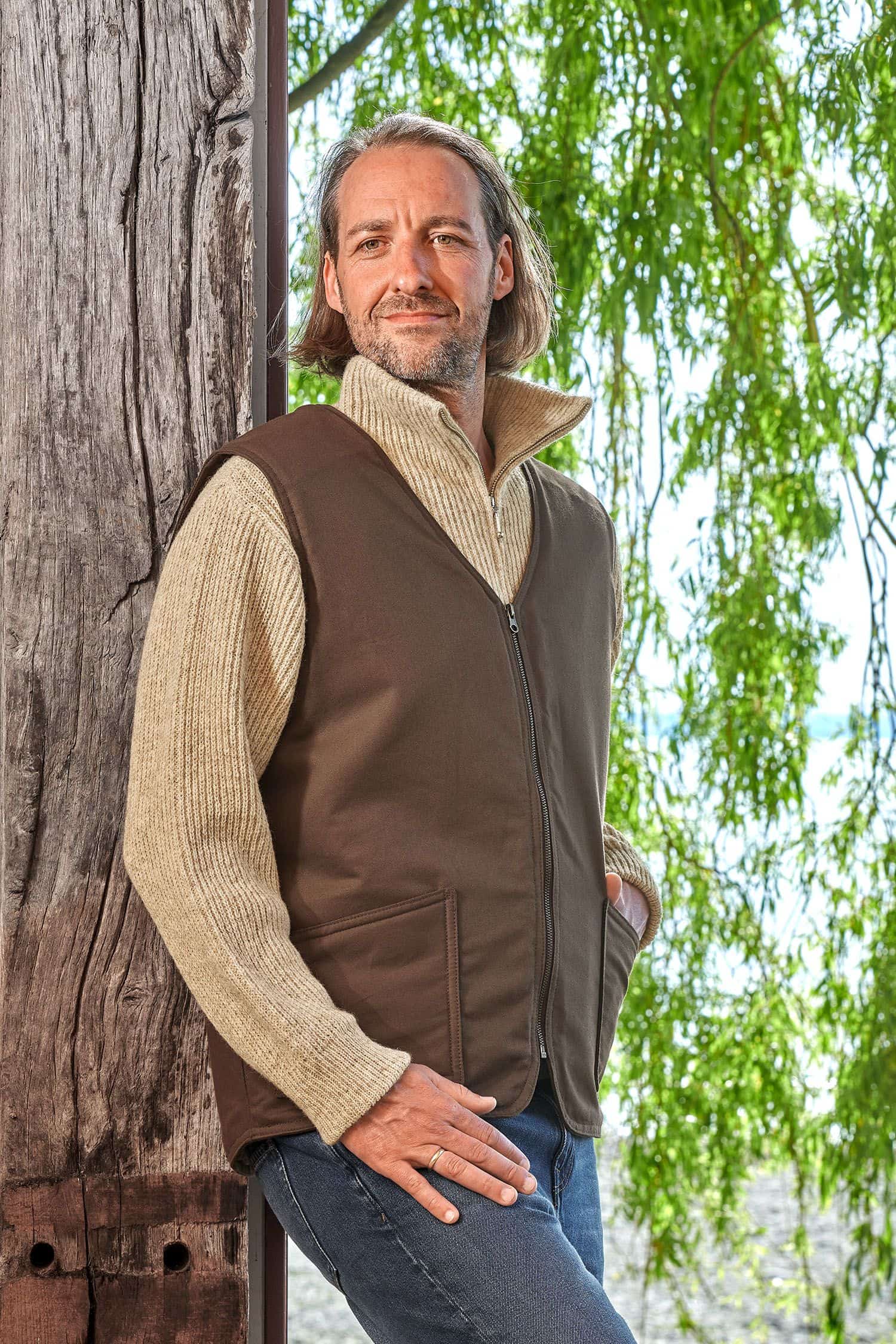 Mann lehnt sich an Baum und trägt einen Troyer und die braune Fellweste aus bio Baumwolle (kbA) und Fell vom Finkhof