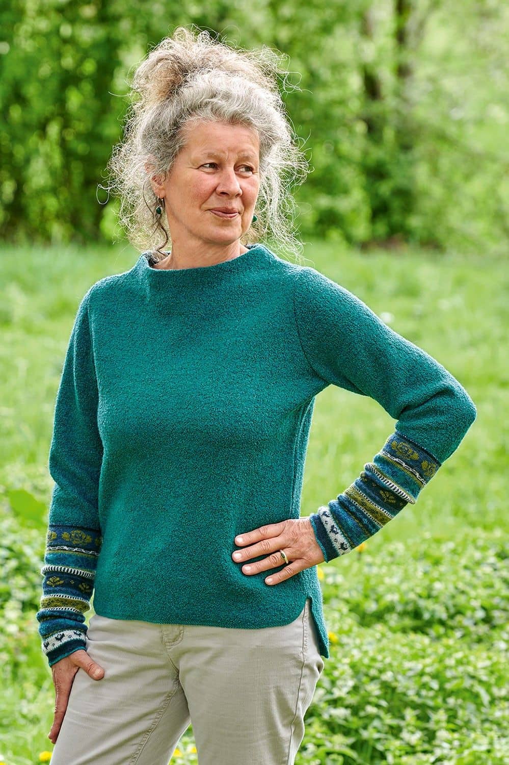 Frau steht in der Natur und trägt türkisen Pulli aus bio Wolle (kbT) und gemusterten Armstulpen
