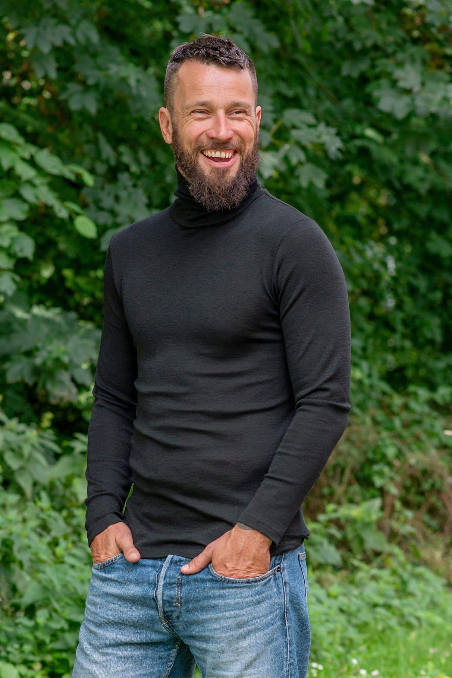 lachender Mann steht im Garten und trägt schwarzen Unterziehrolli aus bio Wolle (kbT) und Seide