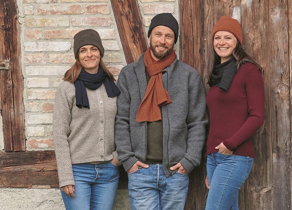 Menschen vor Holztor tragen Strickjacken,Pullis und Schals mit Wollmützen vom Finkhof aus bio Wolle (kbT)