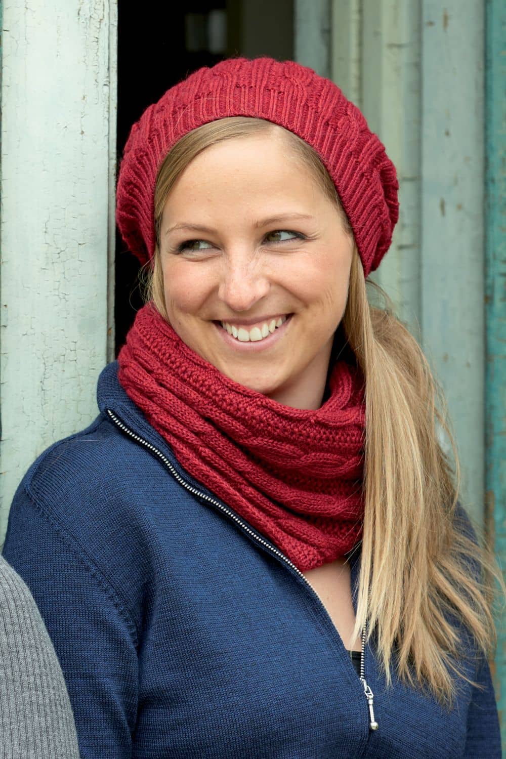 Zopfloop rot Frau trägt blaue Weste, rote Mütze und roter Loopschal mit Zopfmuster aus bio Wolle (kbT) vom Finkhof