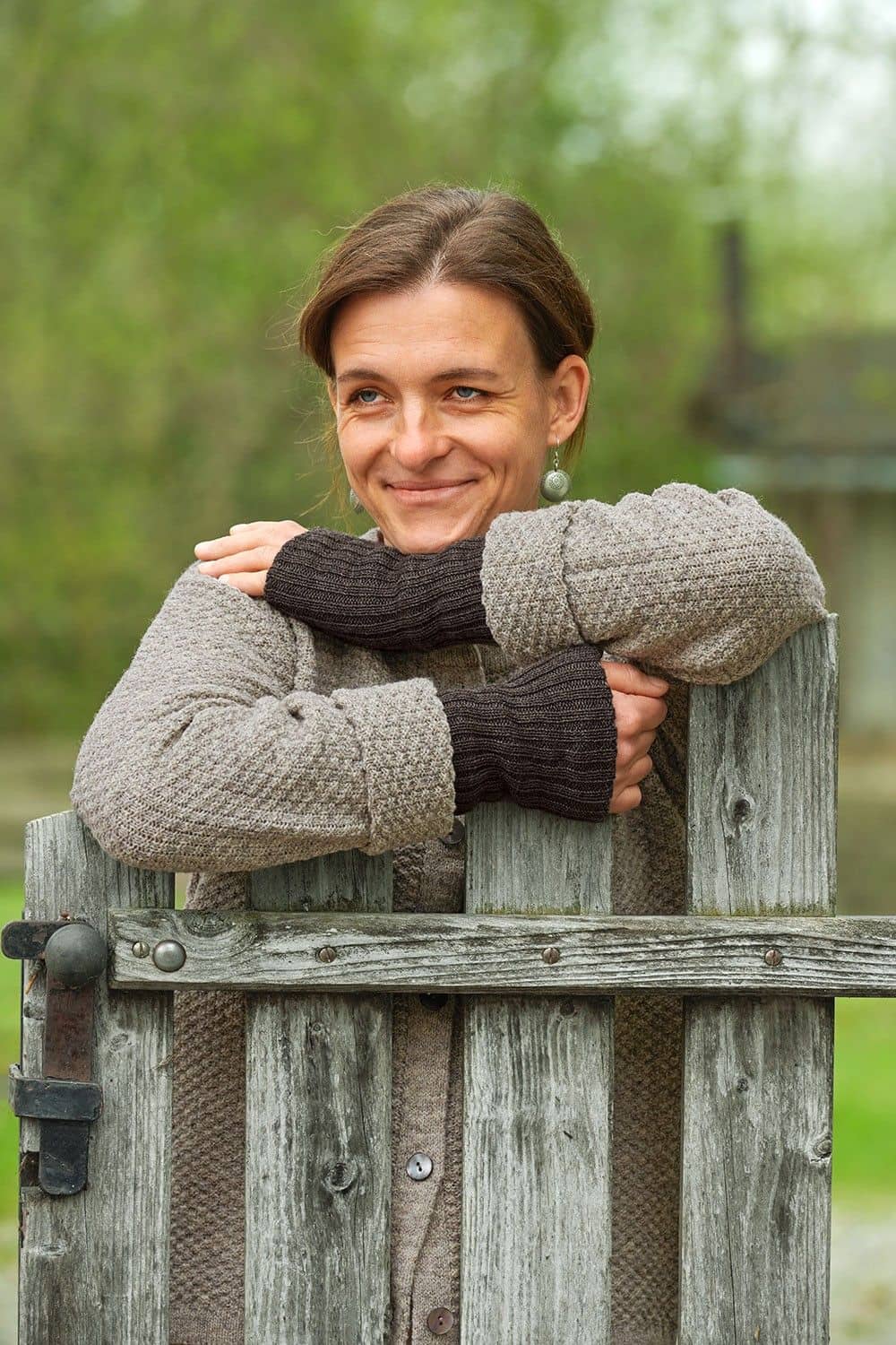 Frau mit grauer Strickjacke und braunen Pulswärmern aus bio Wolle (kbT) vom Finkhof lehnt sich auf Zaun