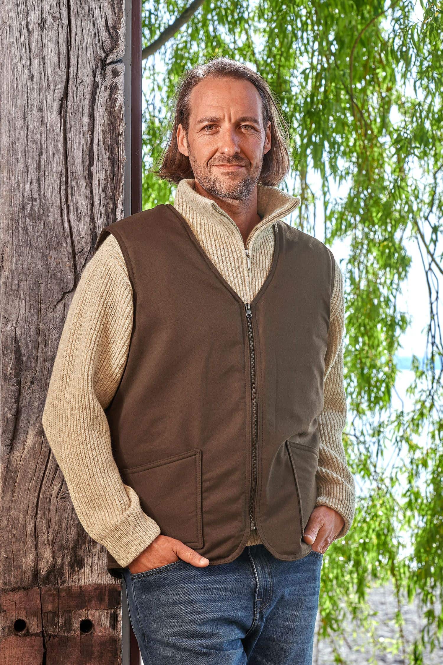 Mann lehnt sich an Baum und trägt einen Troyer und die braune Fellweste aus bio Baumwolle (kbA) und Fell vom Finkhof