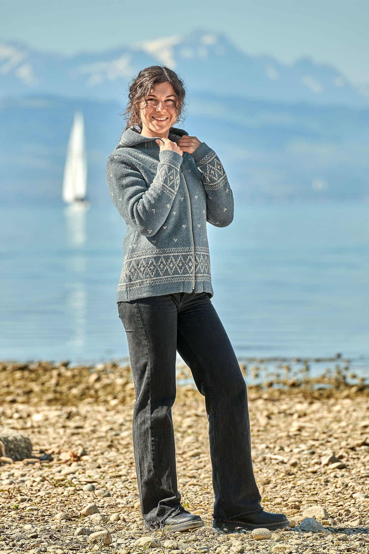 Finkhof-Norweger-Kapuzenjacke Frau vor Bergseekulisse trägt eine rauchblaue Norweger Kapuzenjacke aus bio Wolle (kbT) vom Finkhof und eine schwarze Jeans