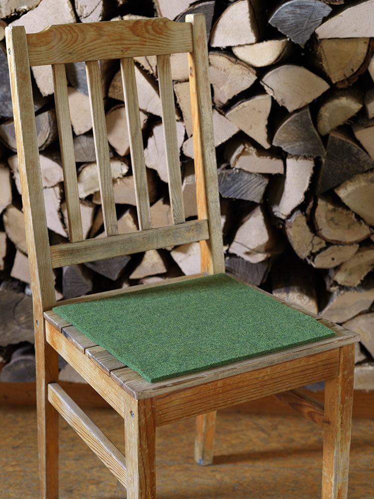 Finkhof-Sitzauflagen aus Nadelfilz quadratisch grün grüne eckige Finkhof Sitzauflage aus bio Wolle (kbT) auf einem Stuhl vor einer Holzwand