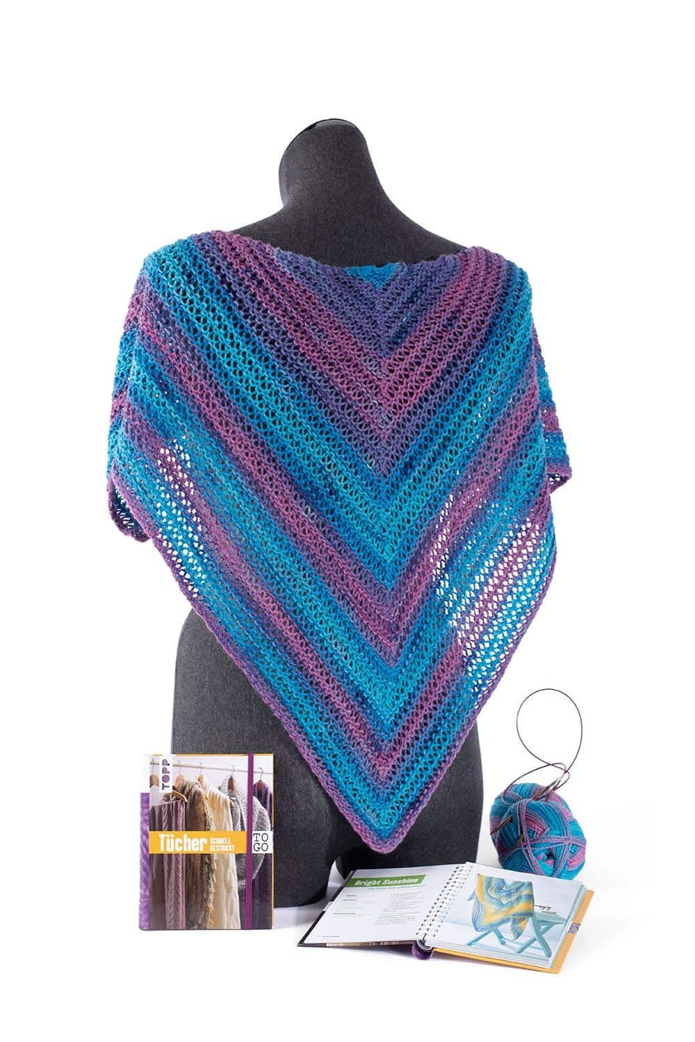 Puppenkörper mit gestricktem Schal mit blau lila Farbverlauf aus der Finkhof Sockenwolle liegt mit Bücher und Wolle