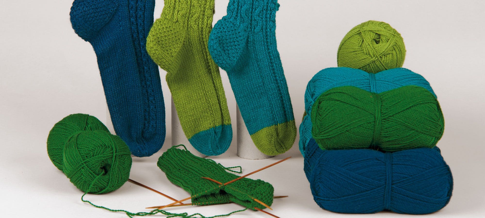 Strickanleitung Socken Finkhof Wolle in grün und blau als Pyramide auf der Seite, gestrickte Socken im Hintergrund und grünem Strickstück davor