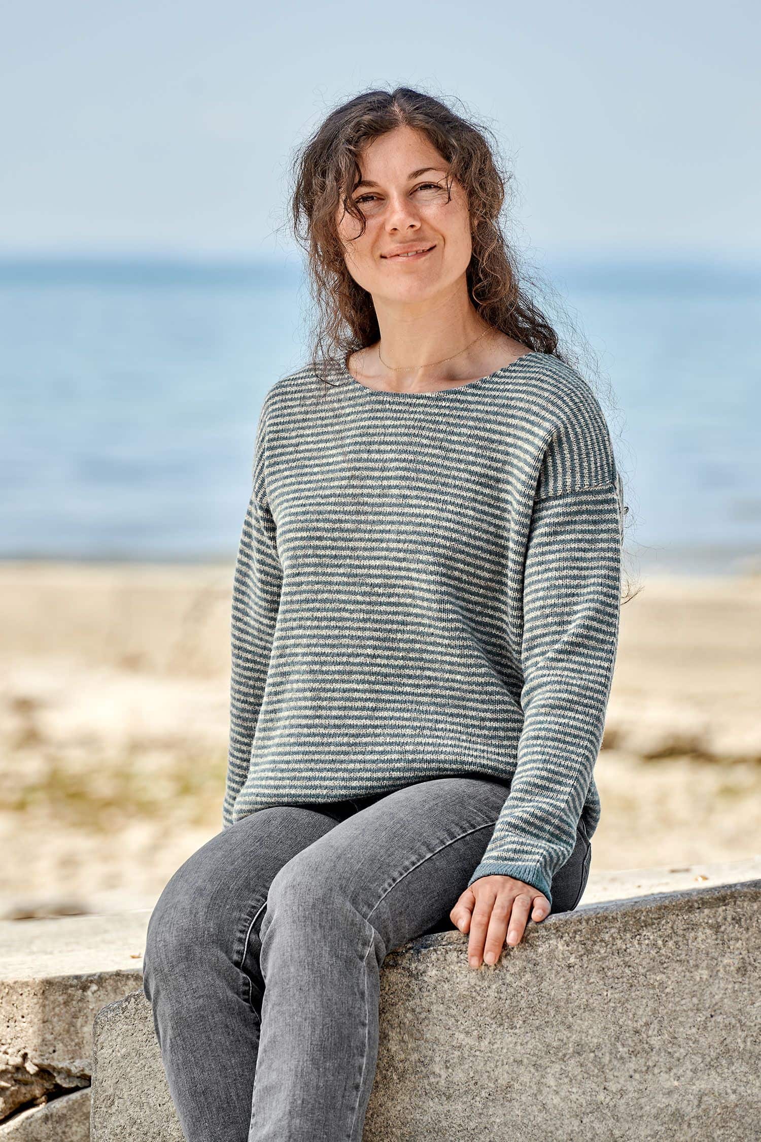 Frau sitzt auf Stein und trägt einen grau blauen Ringelpulli vom Finkhof aus bio Merinowolle (kbT)