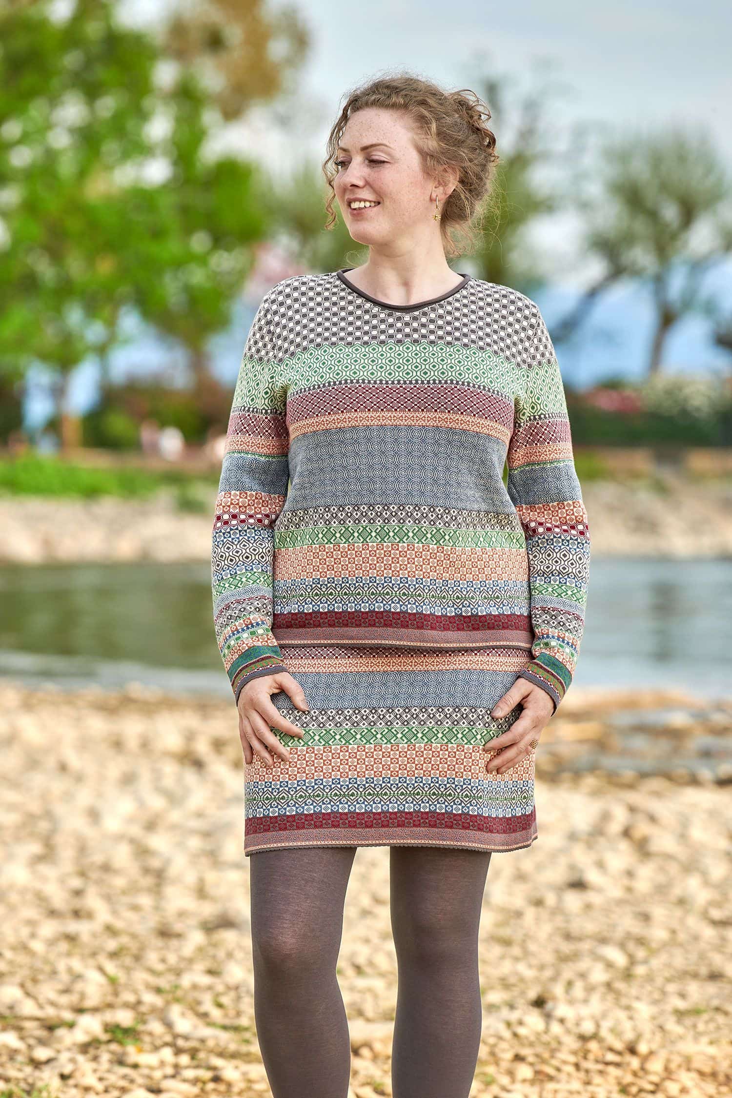 Frau trägt einen gemusterten Rock und Strickpullover aus bio Baumwolle (kbA) und eine lila Strumpfhose
