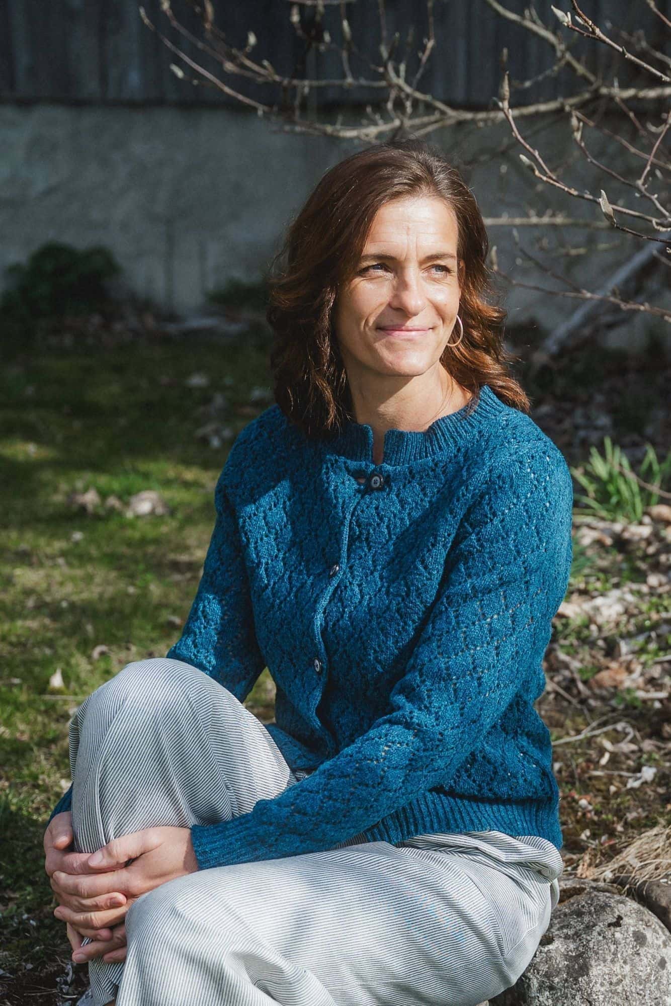 Frau sitzt auf Steinen trägt eine helle Hose mit einer türkisblauen Strickjacke aus Bio-Wolle vom Finkhof