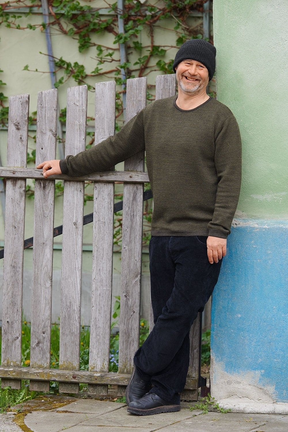 Mann lehnt mit grauer Mütze an Wand und trägt olivegrünen Melangepullover aus bio Wolle (kbT) vom Finkhof