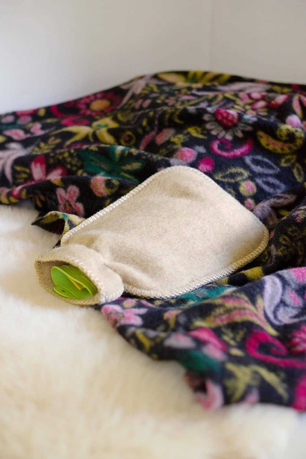 naturweißer Wärmflaschenbezug aus bio Wolle (kbT) vom Finkhof liegt auf Teppich mit floralem Muster