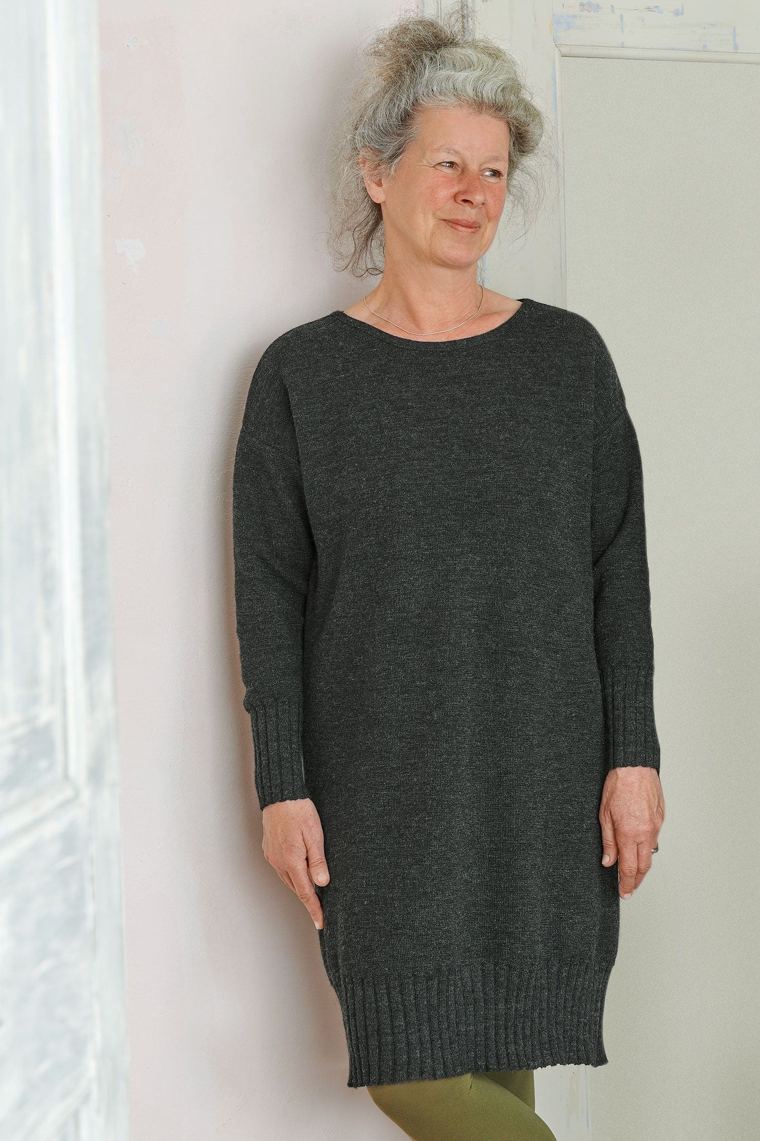 Frau lehnt an Wand und trägt ein dunkelgraues Strickkleid aus bio Wolle (kbT) mit einer grünen Strumpfhose