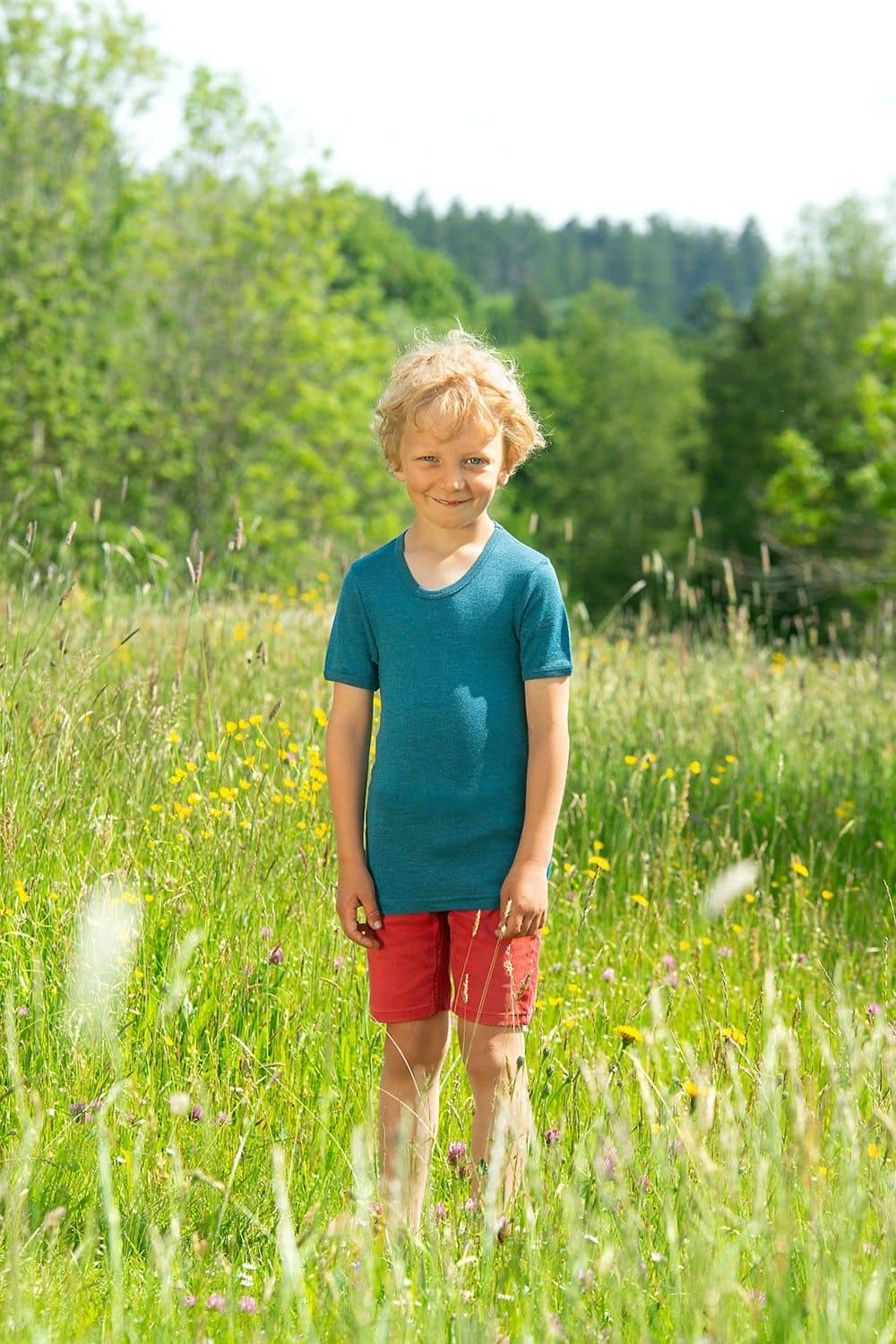Junge steht auf Wiese und trägt rote Hose und türkises Kurzarmshirt aus bio Wolle (kbT) und Seide