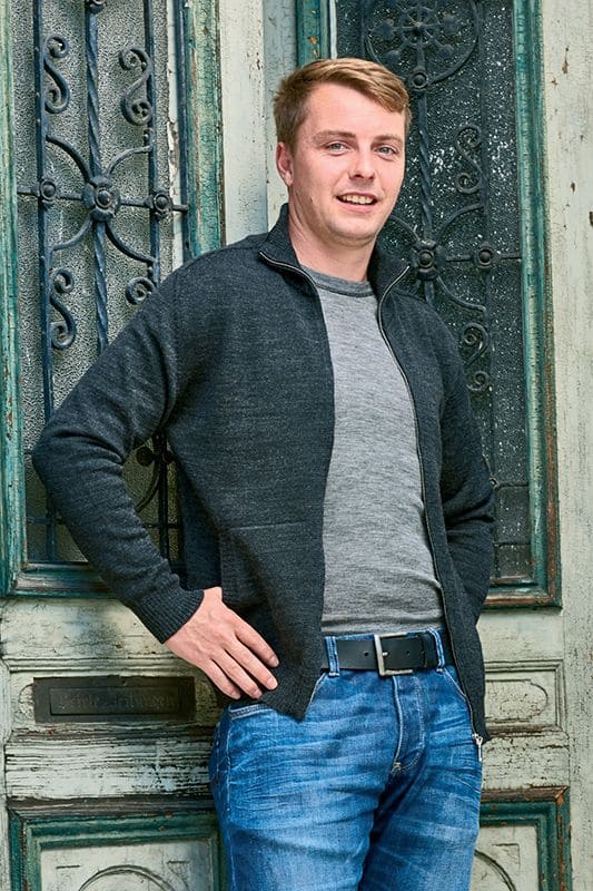 Mann mit grauem Shirt und dunkelgrauer Jacke aus bio Wolle (kbT) lehnt an einer Wand