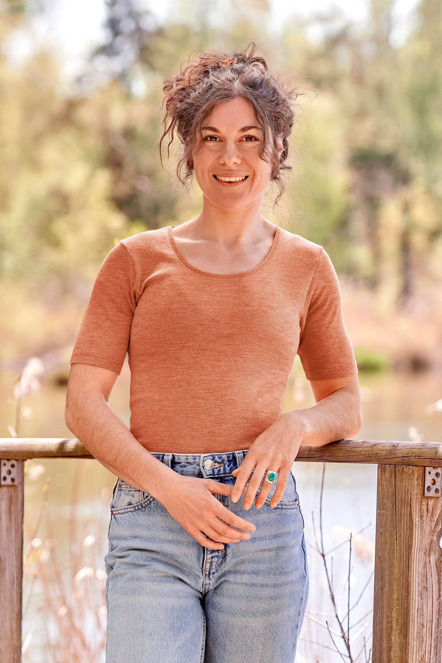 Frau mit Jeans lehnt an Geländer und trägt ein oranges Kurzarmshirt aus bio Wolle (kbT), bio Baumwolle (kbA) und Seide