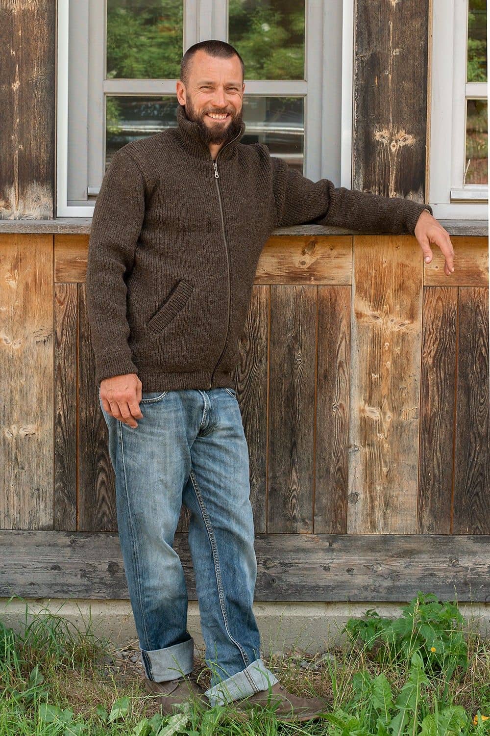 Mann lehnt sich an Holzfassade und trägt Jeans mit brauner Finkhof Jacke aus bio Wolle (kbT)