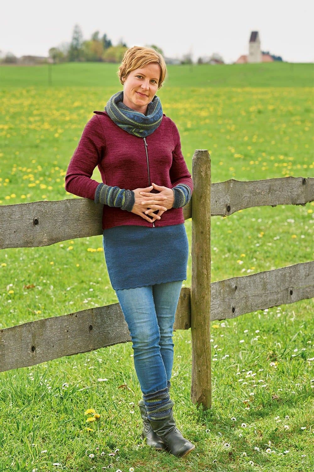 Frau lehnt mit roter Jacke, gemustertem Schal, Stulpen und blauem Kurzrock aus bio Wolle (kbT) vom Finkhof an einem Zaun