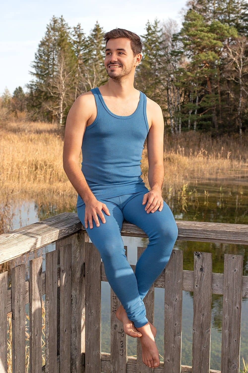 Mann sitzt auf Geländer und trägt blaues Unterhemd aus bio Wolle (kbT) und eine lange Unterhose