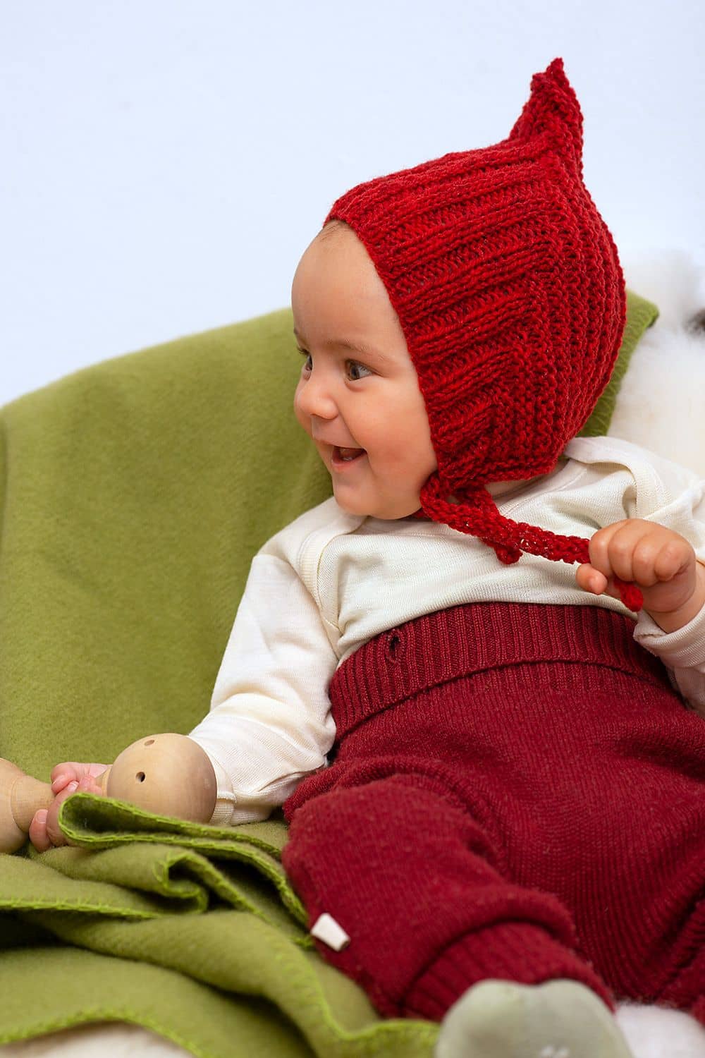 Kind trägt rote Wollhose, weißes Shirt und rote gestrickte Zwergenmütze und sitzt dabei auf einem grünen Teppich