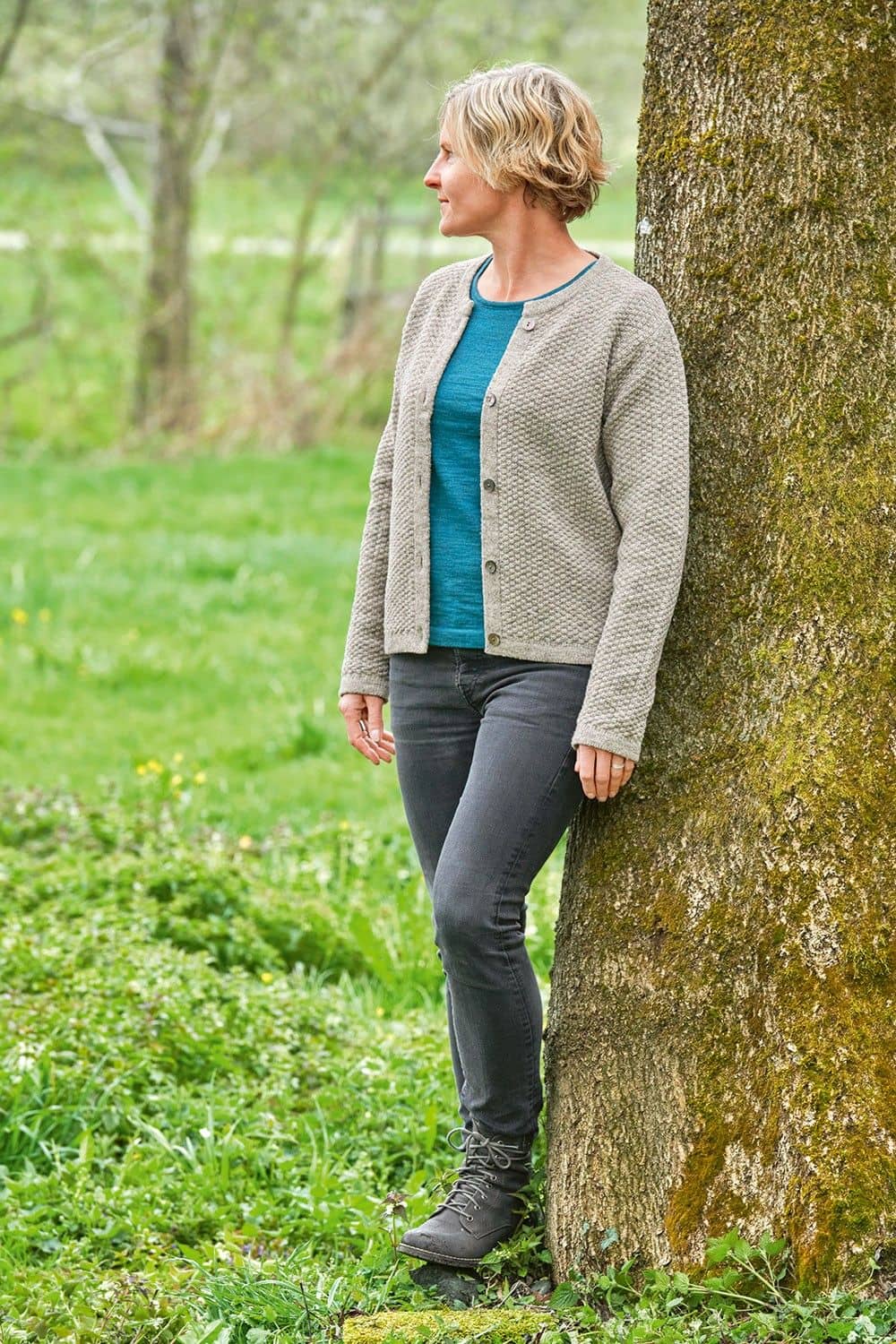 Finkhof-Strukturjacke grau Gr. L Frau lehnt an Baum und trägt graue geöffnete Strukturjacke vom Finkhof aus bio Wolle (kbT) mit türkisem Shirt und Jeans