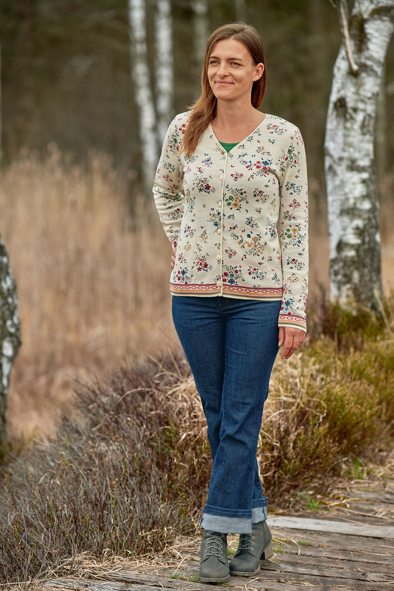 Frau trägt weißen Cardigan mit floralem Muster aus bio Merinowolle (kbT) und bio Baumwolle (kbT) mit Jeans auf einem Steg