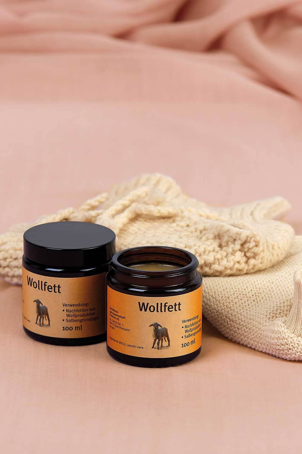 zwei 100ml Glastigel mit Finkhof Wollfett mit Strickwindelhose im Hintergrund, ein Tiegel ist geöffnet