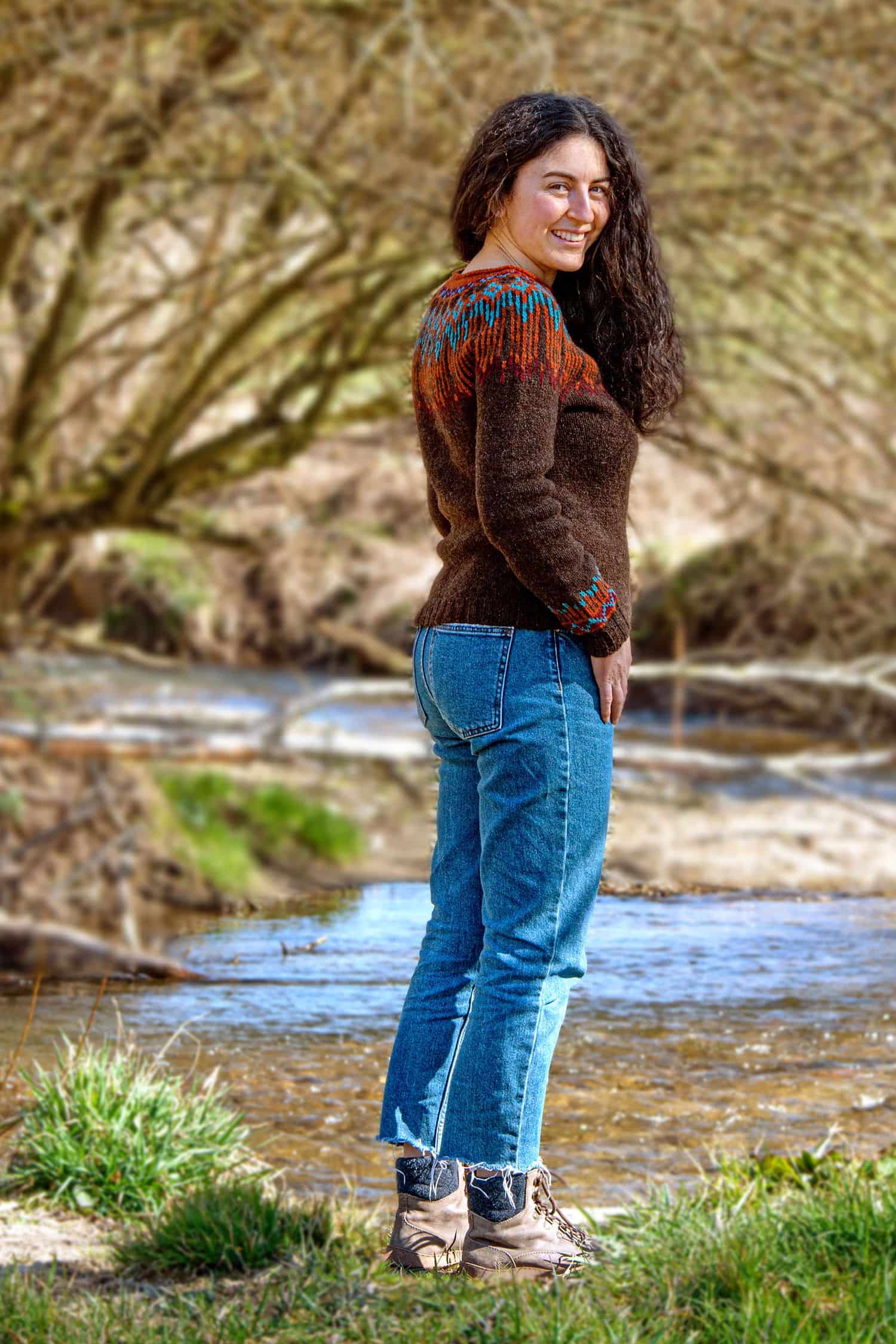 Frau steht neben einem Bach und trägt eine Jeans sowie ein braun gemusterter Pulli aus bio Merinowolle (kbT) vom Finkhof