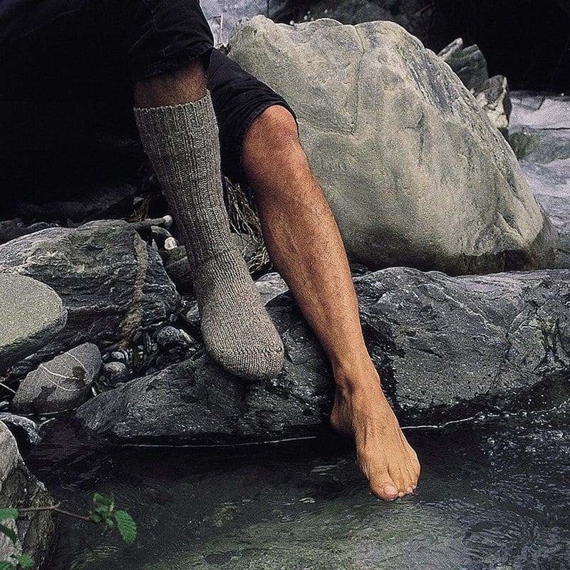 Person trägt an einem Fuß eine graue Wollsocke, den anderen taucht sie in einen Bach