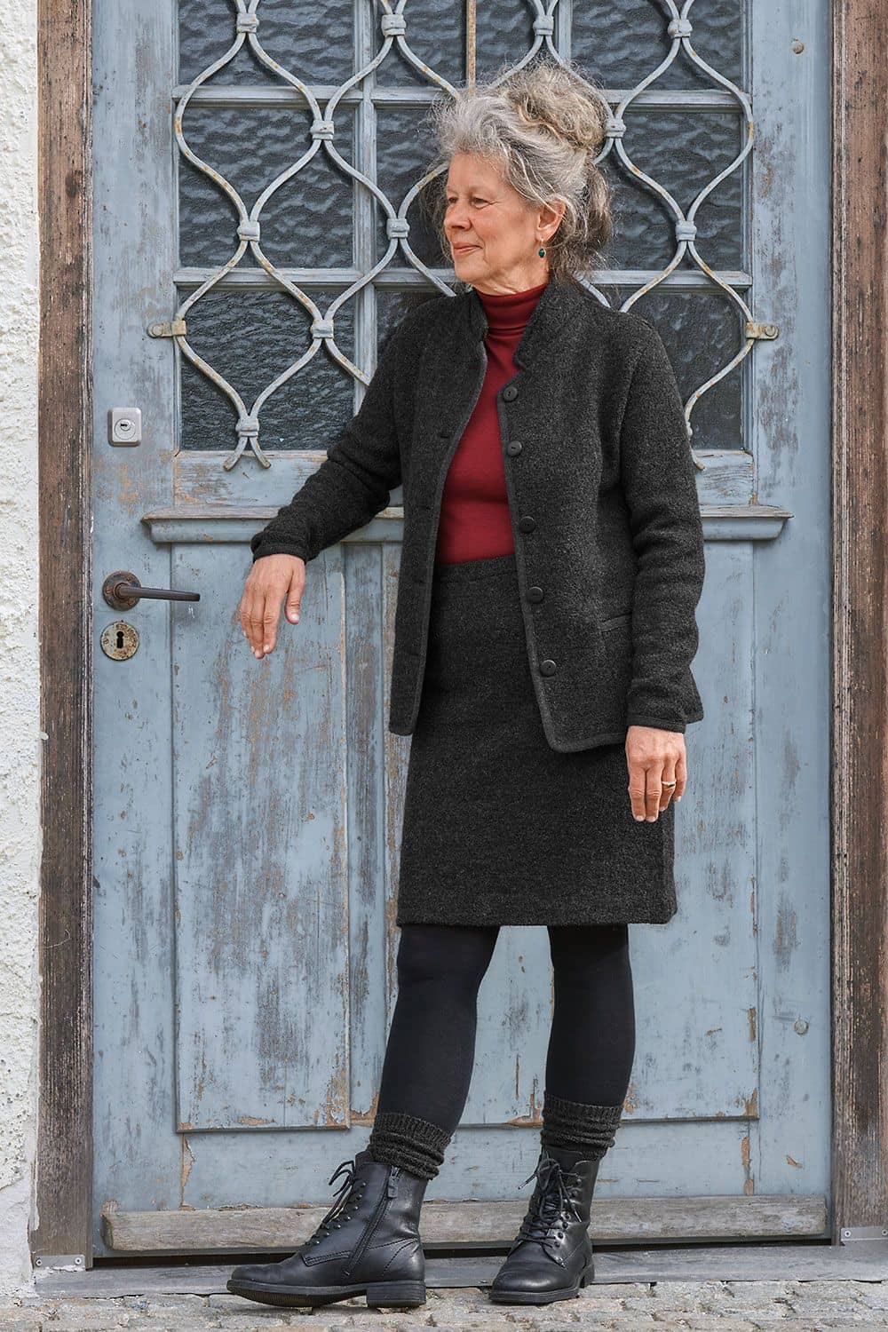 Frau trägt dunkelgrauen Wollrock und Woll Blazer mit rotem Shirt, Stiefeln und Strumpfhose und steht vor einer blauen Tür