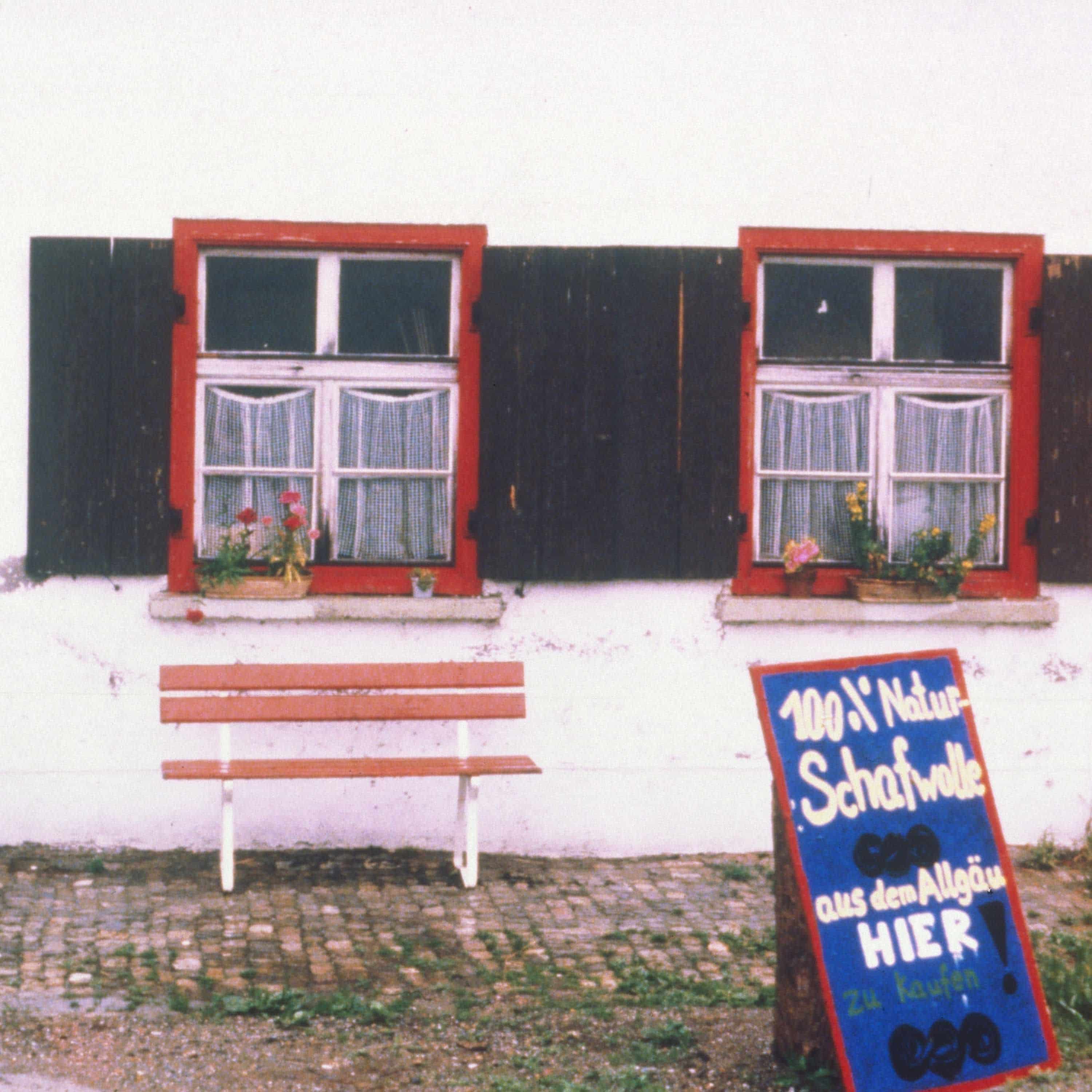 blauer Finkhof Aufsteller vor Haus mit grüenen Fensterläden und einer Bank