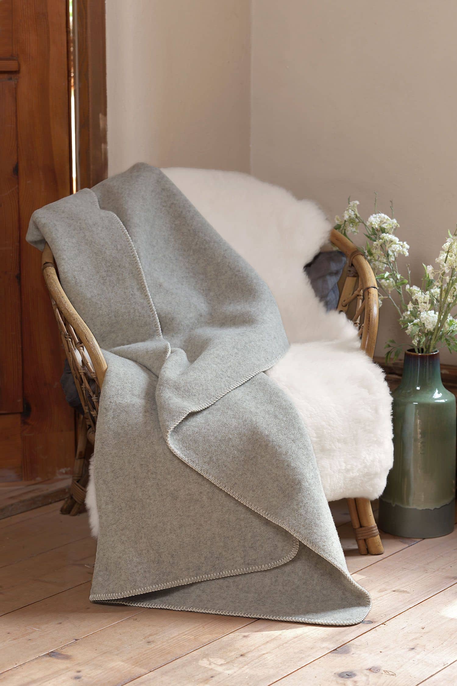 hellgraue Wolldecke aus bio Merinowolle (kbT) vom Finkhof liegt auf Korbstuhl mit weißem Fell