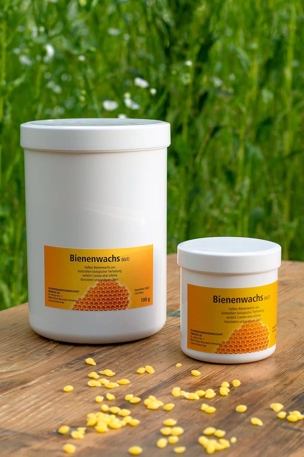 Finkhof Bienenwachs (kbA) in weißen 100g und 500g Dosen mit Wachsdrops davor, Wiese im Hintergrund