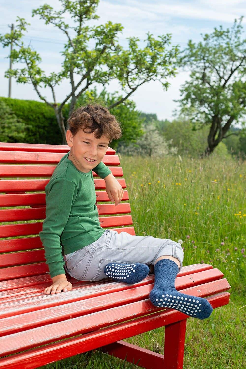 Kind sitzt auf roter Bank und trägt ein grünes Shirt und blaue Wollplüsch Stoppersocken aus bio Wolle (kbT)
