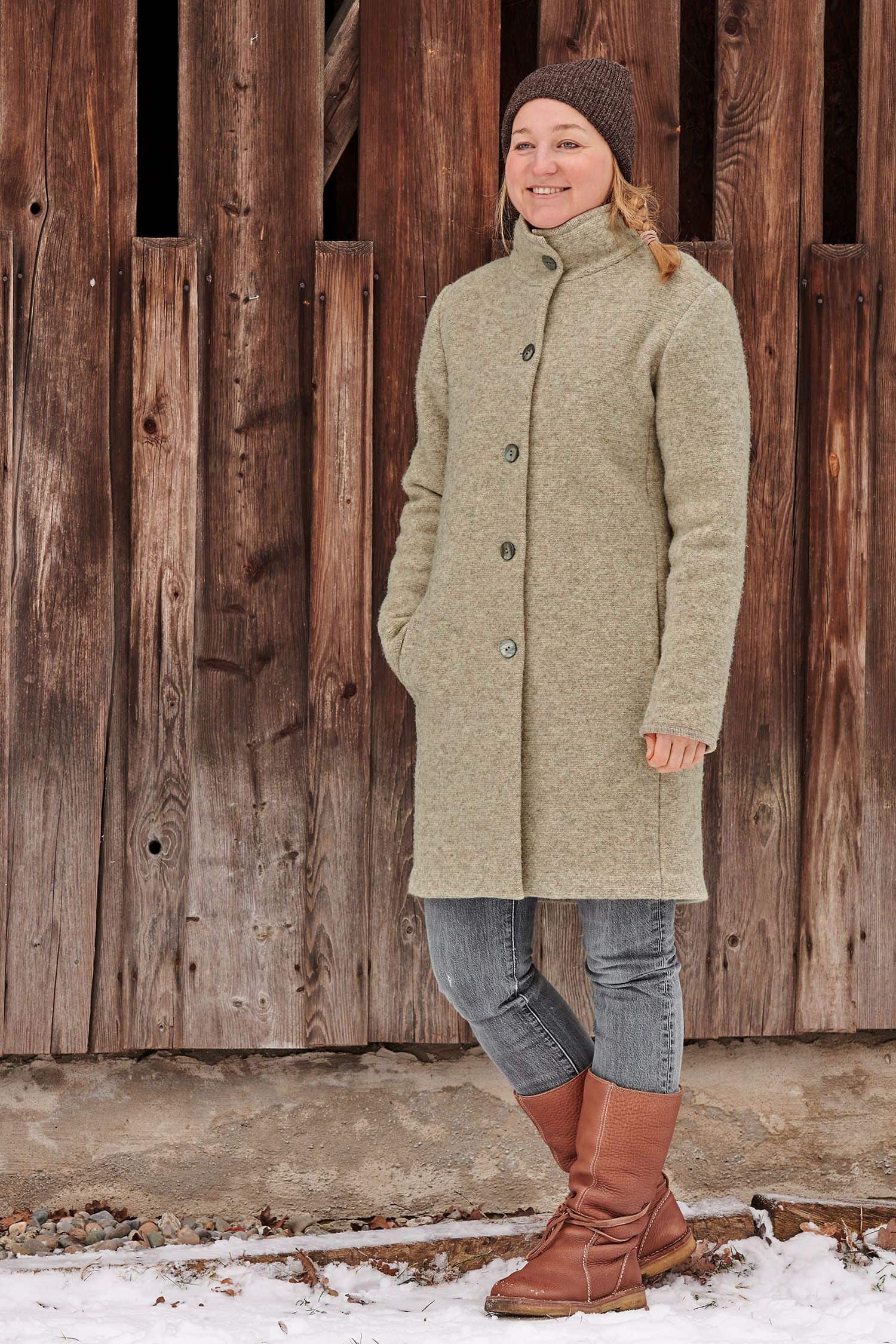 Frau steht vor Holzwand und trägt braune Mütze und einen grauen Finkhof Wollwalkmantel aus bio Wolle (kbT)